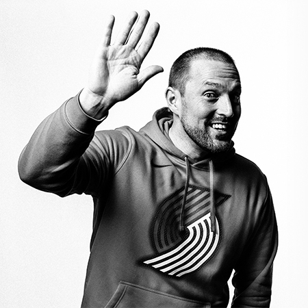 Black and white photo of a man smiling wearing a Blazers sweatshirt and holding his hand up giving a high 5 gesture.