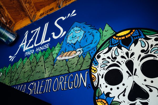 Photo of a mural featuring the text Azuls Taco House, Salem, Oregon with evergreen trees, a blue bigfoot holding tacos and a portion of a sugar skull on a blue background