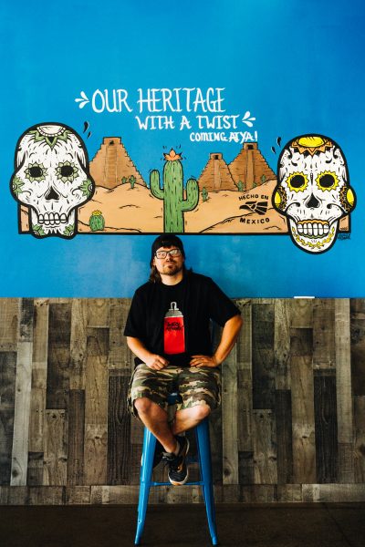 A man wearing a backwards cap and glasses sits on a stool in front of a mural of two sugar skulls and a desert and cactus setting with the text "Our Heritage with a Twist"